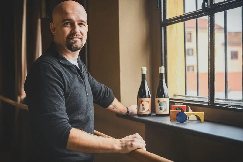 Andrea Moser, créateur du projet AMProject, posant près d’une fenêtre avec deux bouteilles de vin Temporary Wine et des objets design colorés