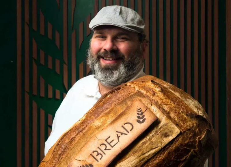 Mario D'Oria, maître boulanger à Alberobello, posant avec une miche de pain artisanal portant l’inscription 'Bread', symbole de son savoir-faire unique chez Profumi dal Ponte.
