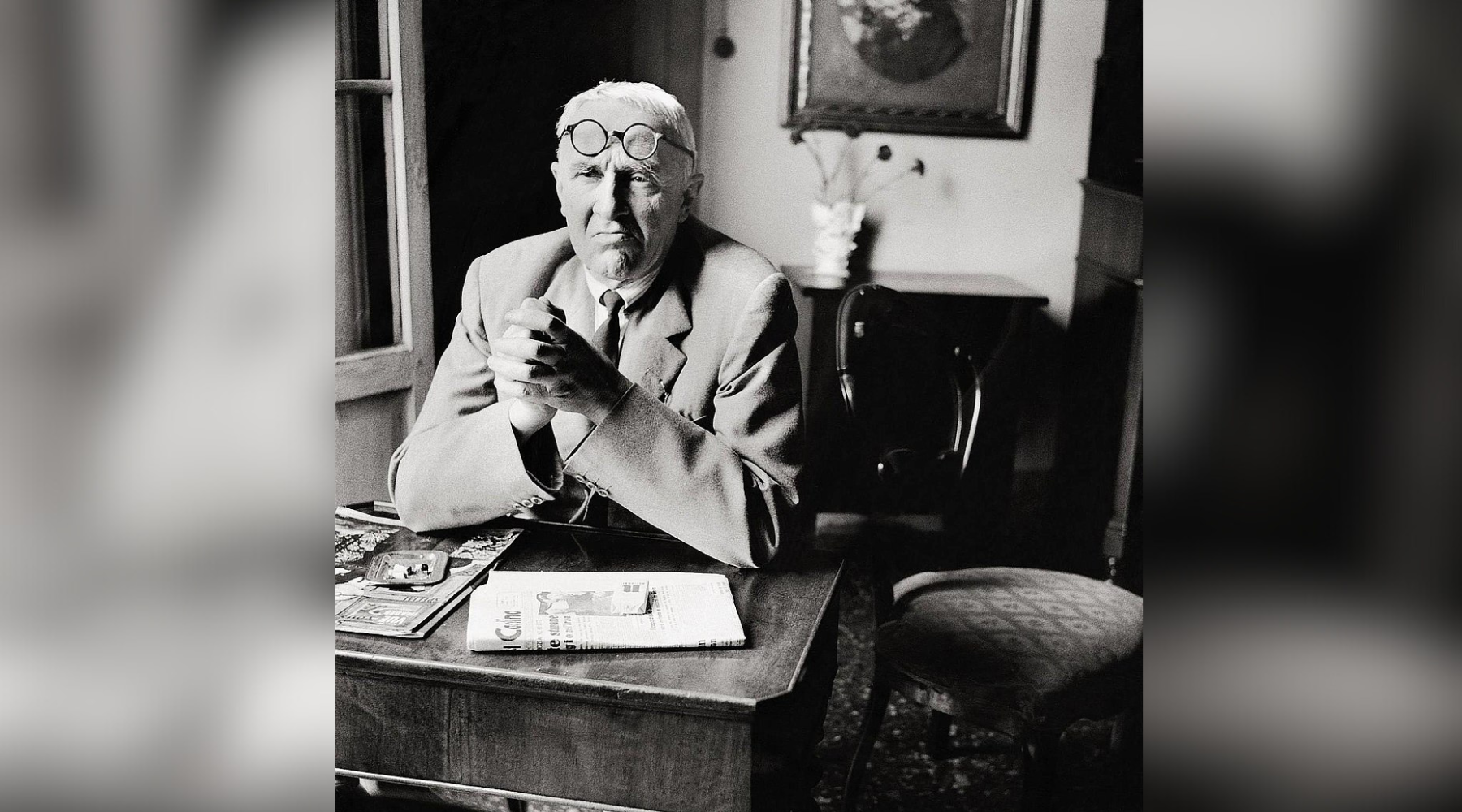 Photographie en noir et blanc du peintre et graveur italien Giorgio Morandi, assis à une table avec des journaux et des magazines. Il porte un costume et des lunettes rondes posées sur son front, dans un intérieur au style classique.