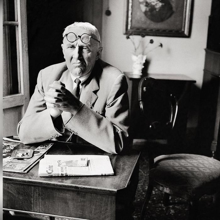 Photographie en noir et blanc du peintre et graveur italien Giorgio Morandi, assis à une table avec des journaux et des magazines. Il porte un costume et des lunettes rondes posées sur son front, dans un intérieur au style classique.