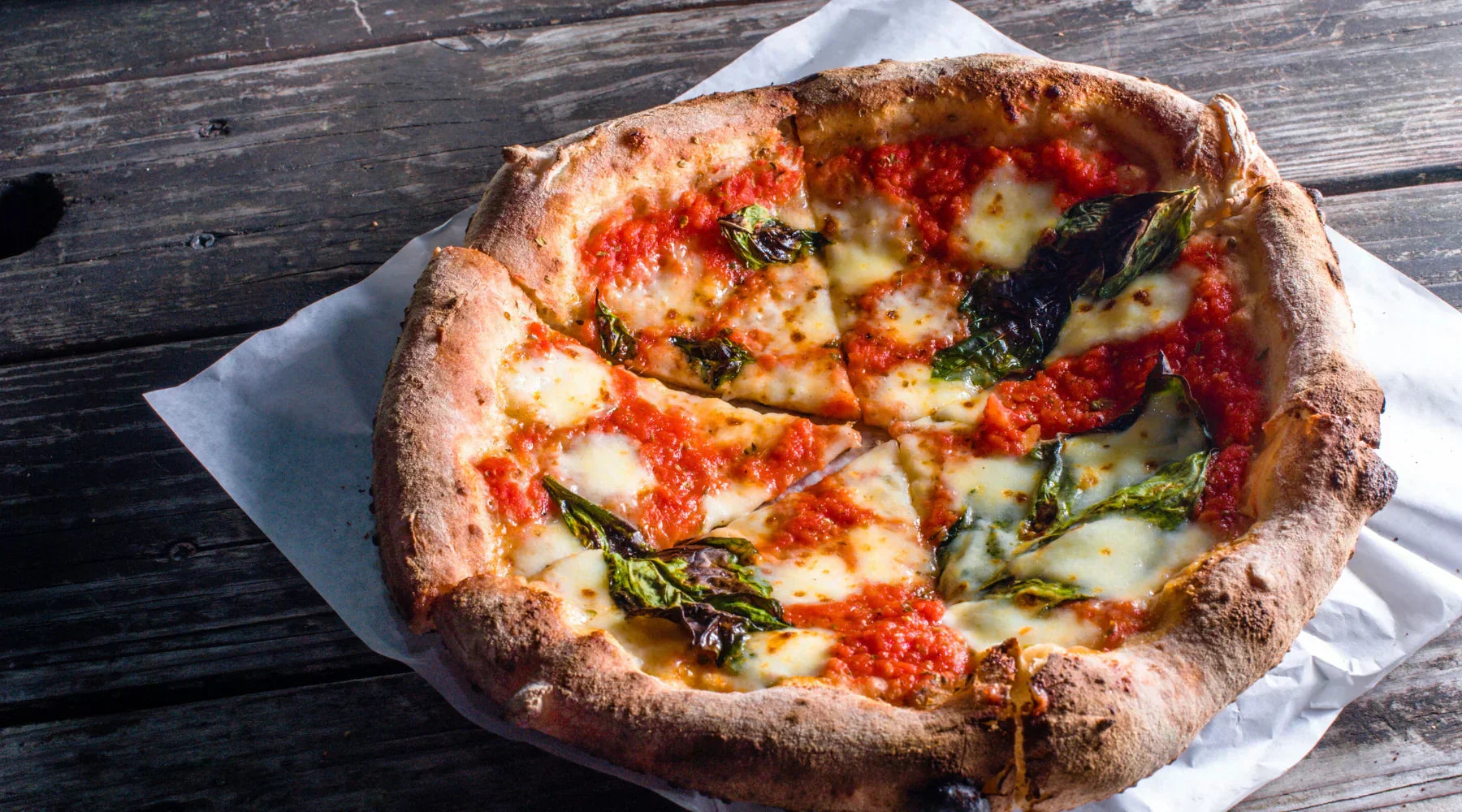 Pizza Margherita artisanale à la croûte dorée et gonflée, garnie de mozzarella fondante, de tomates San Marzano et de basilic frais, servie sur une table rustique en bois. Symbole de la tradition napolitaine et de l’authenticité italienne.