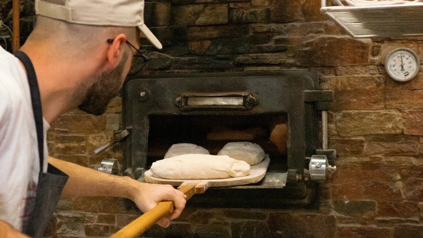 Un boulanger enfournant du pain artisanal dans un four traditionnel en pierre chez Eataly Smeraldo, illustrant le processus de cuisson du pain au levain.