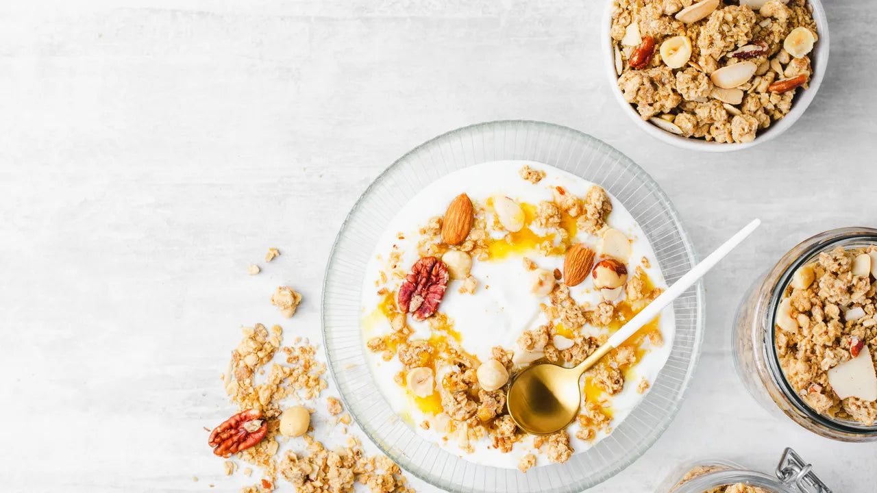 Bol de yaourt grec garni de granola, noix et miel, présenté avec des ingrédients en accompagnement sur fond clair