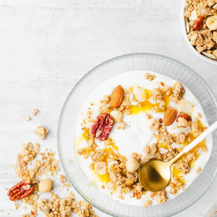 Bol de yaourt grec garni de granola, noix et miel, présenté avec des ingrédients en accompagnement sur fond clair