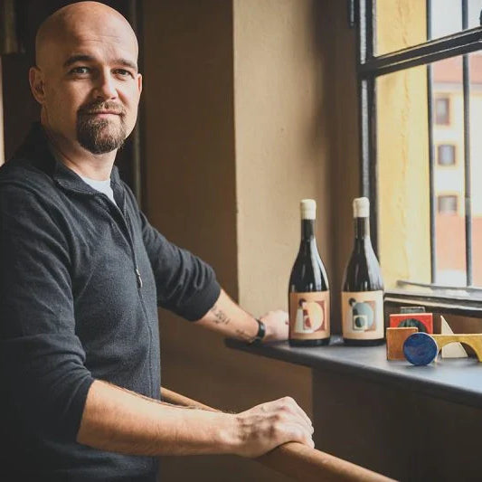 Andrea Moser, créateur du projet AMProject, posant près d’une fenêtre avec deux bouteilles de vin Temporary Wine et des objets design colorés