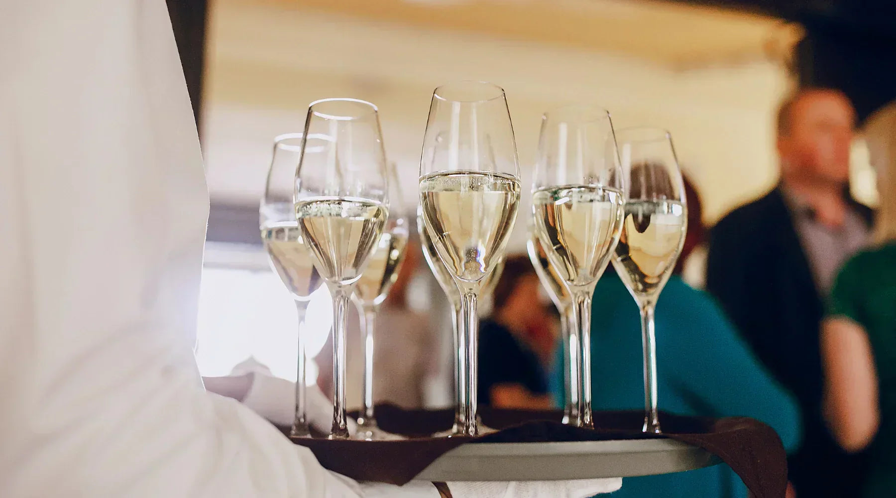 Serveur en tenue élégante portant un plateau de coupes de champagne lors d'un événement raffiné, avec des invités en arrière-plan dans une ambiance festive.